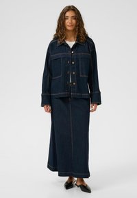 Dunkelblaue Jeansjacke mit Brusttaschen und Knöpfen, kombiniert mit einem passenden langen Jeansrock. Das Outfit verfügt über sichtbare Nähte.