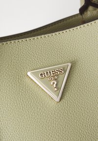 Fényes zöld textúrájú bőrtáska, amelyen egy háromszög alakú, aranyozott logó található "GUESS USA" felirattal és kérdőjel részlettel.