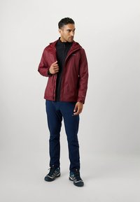 Vinröd vattentät jacka med huva, svart dragkedjejacka under, kombinerad med blå byxor och gråblå sneakers, stående mot en neutral bakgrund.