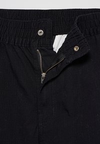 Pantalon noir avec taille élastique, fermeture éclair métallique partiellement ouverte et deux boutons-pression métalliques, montrant la doublure intérieure blanche.