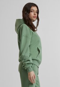 Sweat à capuche vert avec une coupe ample, en tissu doux, poche kangourou, poignets côtelés et capuche doublée. Porté avec un pantalon assorti.