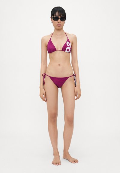 Ensemble bikini en magenta vif avec un motif et un logo sur le haut. Présente des bonnets triangulaires et un bas à nouer sur les côtés. Des lunettes de soleil complètent le look.