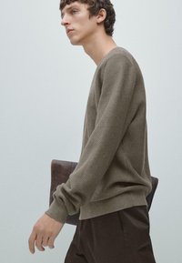 Olivgrüner, strukturierter Pullover mit geripptem Saum, getragen mit dunkelbraunen Hosen, vor einem hellgrauen Hintergrund gehalten.