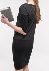 Das schwarze Kleid hat einen glatten, engen Rock und ein lockeres, kurzärmeliges Oberteil mit einem durchsichtigen Rückenteil. In der Hand hält sie eine glitzernde, dunkle Clutch.