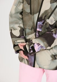 Jacke im Camouflage-Muster in gedämpften Grün- und Violetttönen, mit Reißverschluss, verstellbaren Manschetten und einem Etikett mit der Aufschrift "W-PRO 10000".