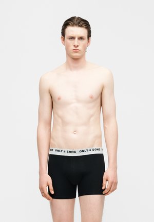 Młody mężczyzna o jasnej karnacji i brązowych włosach, ubrany w czarne bokserki z białym pasem z napisem "ONLY & SONS", stoi na jednolitym tle.