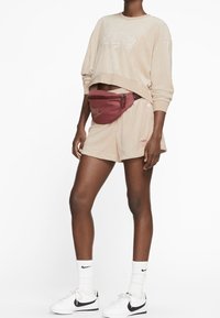 Beige kortärmad sweatshirt med vitt Nike-logga, ihopparad med beiga shorts. Röd midjeväska med guldlogga, vita sneakers och strumpor i ankellängd.