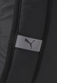 Tessuto nero con una texture liscia presenta una striscia grigia contenente un logo Puma nero. Il design è minimalista e atletico.