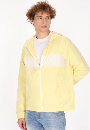 Chaqueta amarilla con cremallera y una franjas horizontal blanca, hecha de material ligero, con capucha y bolsillos laterales, combinada con jeans azules.