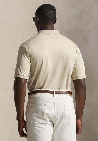 Homme portant un polo beige à manches courtes, pantalon blanc avec ceinture marron, lunettes de soleil et montre, debout, dos tourné vers le spectateur.