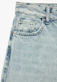 Jeans de mezclilla azul claro con una textura desteñida, que cuentan con un bolsillo frontal, costuras prominentes y un acento de remache de cobre en la cinturilla.