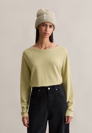 Lichtgroen langemouwen top met ribbelstructuur, gecombineerd met hoge taille zwarte broeken. Het model draagt een beige gebreide beanie.