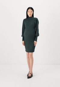 Robe en tricot vert foncé avec un col montant, texture côtelée et manches longues bouffantes. Associée à des chaussures à talons noires, silhouette ajustée.