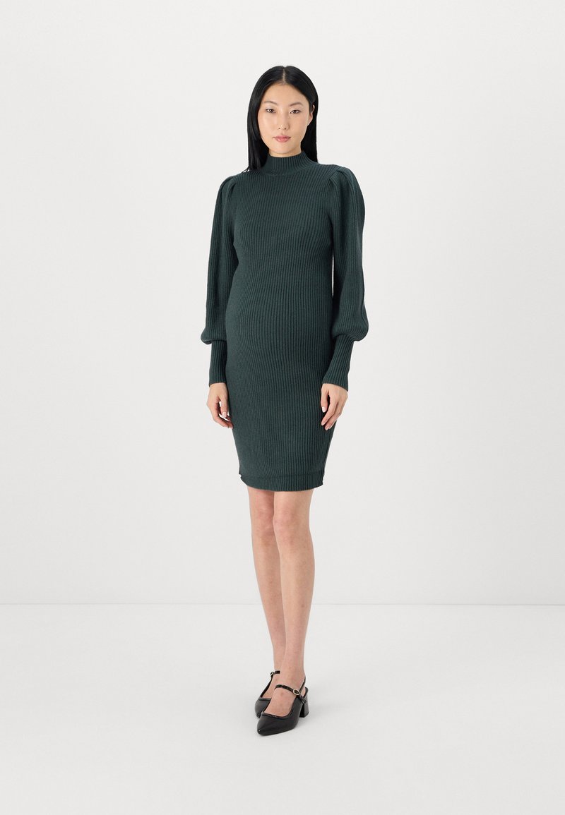 Robe en tricot vert foncé avec un col montant, texture côtelée et manches longues bouffantes. Associée à des chaussures à talons noires, silhouette ajustée.