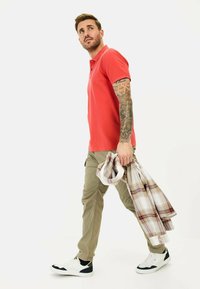 Rød polo-shirt, khaki cargo-bukser, bærende en ternet skjorte. Hvide og sorte sneakers. Tatoveringer på armen. Neutral baggrund.