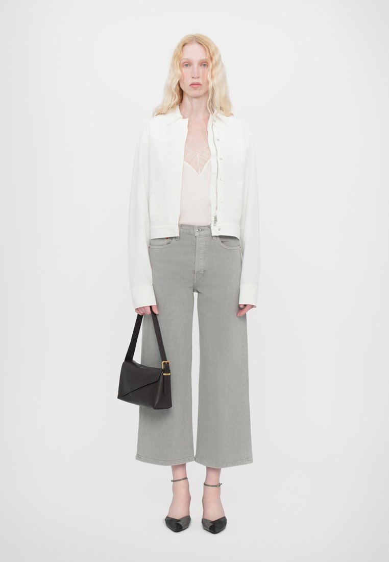 Veste blanche raccourcie, haut léger transparent, jeans gris à jambes larges, sac à main noir et chaussures à talons noires ; tissu lisse, design simple.