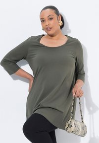 Vihreä oliivinvärinen tunika, jossa on pyöreä kaula-aukko ja kolmannes-hihat, yhdistetty mustiin legginsseihin. Pitelee pientä, beigeä käärmeennahkakuosisia käsilaukkua.