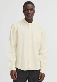 Camisa de manga larga, color crema, con cuello mao, bolsillo frontal y cierre de botones. La tela parece suave, ligera y lisa.