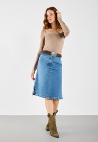 Jeanskjol med en framspring, kombinerad med en tan långärmad tröja och ett flätat brunt bälte. Modellen bär olivgröna ankelskor.