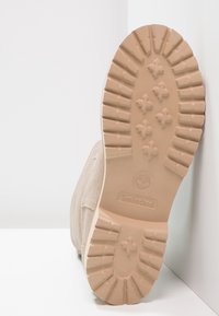 Béžová semišová bota s tlustou, strukturovanou gumovou podrážkou s vzorovým treadem a vtisknutým brandingem. Pohled na podrážku na bílém pozadí.