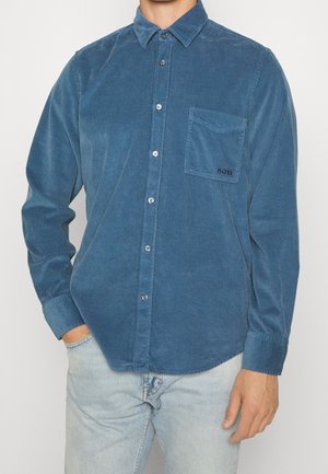 Mies, jolla on päällään sininen mannapintainen napillinen paita, jossa on rintatasku, jossa lukee "BOSS", ja vaaleansiniset farkut, seisomassa yhdellä kädellä taskussa.