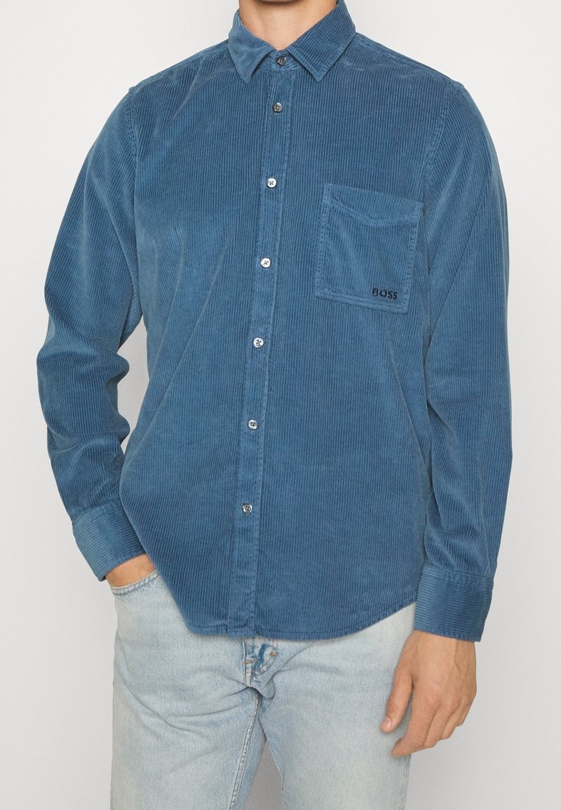 Homme portant une chemise en velours côtelé bleu avec une poche poitrine étiquetée "BOSS" et un jean bleu clair, debout avec une main dans la poche.