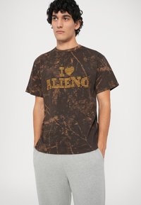 Camiseta marrón con estampado de tie-dye y gráfico amarillo "I ♥ ALIEN", con cuello redondo, mangas cortas y corte relajado, combinada con pantalones de chándal grises.