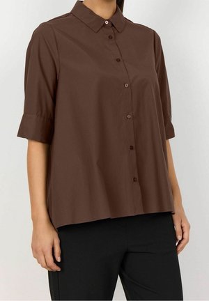 Femme portant une chemise marron ample boutonnée avec des manches jusqu'au coude et un pantalon noir, debout devant un fond clair uni.