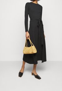 Abito lungo nero a maniche lunghe con cintura legata, lunghezza fino alla caviglia, in tessuto liscio; il modello tiene una piccola borsa intrecciata dorata e indossa mules neri con tacco.