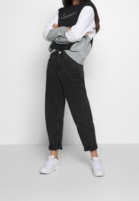 Person standing with arms crossed wearing a black, gray, and white Nike sweatshirt, black jeans, and white sneakers against a plain background.