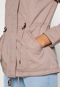 Light pink, fabric jacket with a drawstring waist, flap pockets, and a slightly textured surface. Features a hood with adjustable ties.