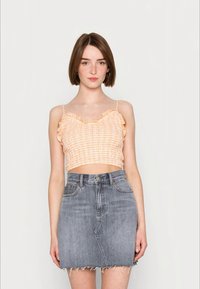Jovem mulher vestindo um top cropped com babados azul xadrez e uma saia de denim cinza de cintura alta com a barra desfiada, de pé contra um fundo simples.