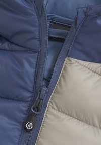 Giacca a vento leggera con esterno blu navy, dettagli beige, chiusura con zip e texture trapuntata per l'isolamento.