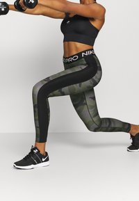 Zwarte sportbh, groene camouflage legging met zwarte zijpanelen en tailleband, en zwarte sport schoenen. Benen in een lungespositie, gewichten in de handen.