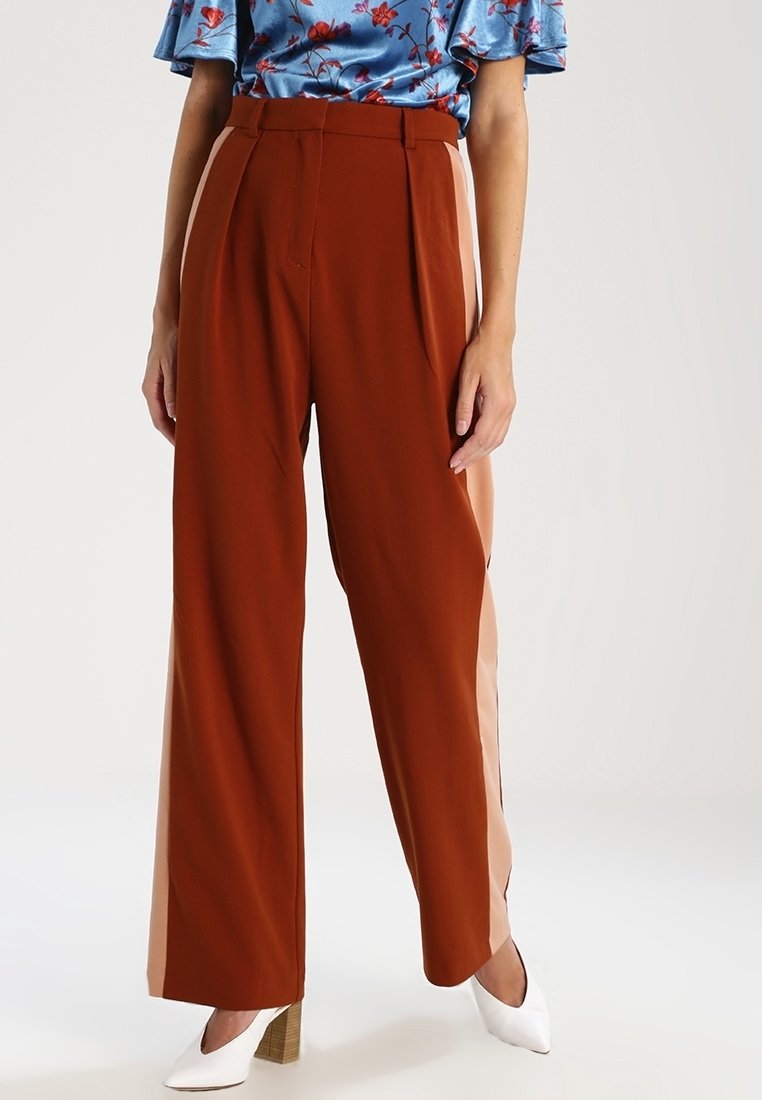 Femme portant un pantalon large couleur rouille avec des bandes latérales beiges, associé à des chaussures blanches à talons et un haut fleuri bleu.