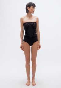 Γυναίκα όρθια ξυπόλητη με μαγιό χωρίς τιράντες μαύρου χρώματος, με διαμαντοειδές σχέδιο μπροστά, κοιτάζοντας προς τα αριστερά της, με φόντο απλό λευκό.