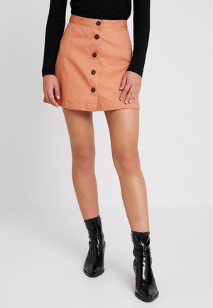 Mujer vistiendo una mini falda color melocotón con botones delanteros, top negro de manga larga y botas negras brillantes hasta el tobillo, de pie sobre un fondo blanco.