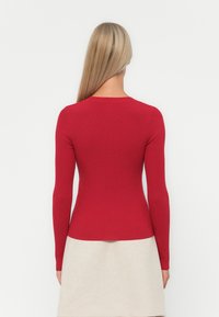 Rød, stripete langermet topp med et tettsittende design, med rund halslinning og teksturert stoff. Brukt sammen med en lysfarget skjørt.
