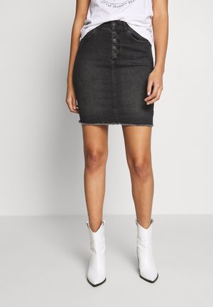 Woman wearing a black buttoned denim skirt and white cowboy boots, standing against a plain gray background.