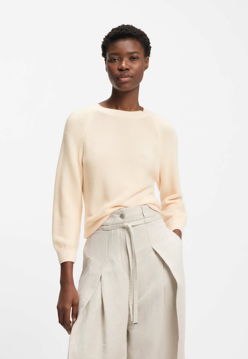 Kvinna med kort lockigt hår bär en ljus persikofärgad tröja och högmidjade beige byxor med ett snörbälte på en vit bakgrund.