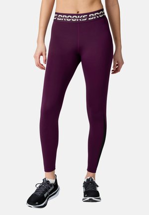 Kobiecy tułów i nogi w ciemnofioletowych legginsach sportowych z markowym pasem i czarnych butach do biegania na jednolitym jasnym tle.