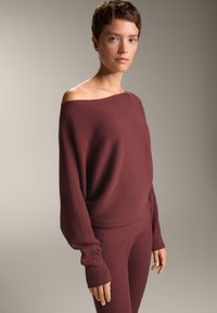 Burgundska rebrasta jopica z off-shoulder dizajnom, dolgimi rokavi in usklajenimi rebrastimi hlačami, ki prikazujejo mehak tekstur in sproščen kroj.