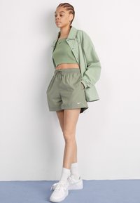 Zelená športová bunda, zodpovedajúci tielko a šortky s logom Nike. Biely tenisky a ponožky dopĺňajú outfit a zdôrazňujú aktívny štýl.