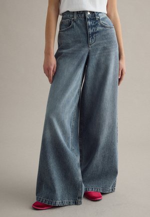 Persona che indossa jeans blu a vita alta e gamba larga abbinati a scarpe piatte rosa, in piedi contro uno sfondo neutro e semplice.