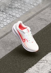Zapatilla deportiva blanca con un acabado texturizado, acentos en rosa y rojo, suela acolchada y diseño con cordones, sobre un fondo de concreto.