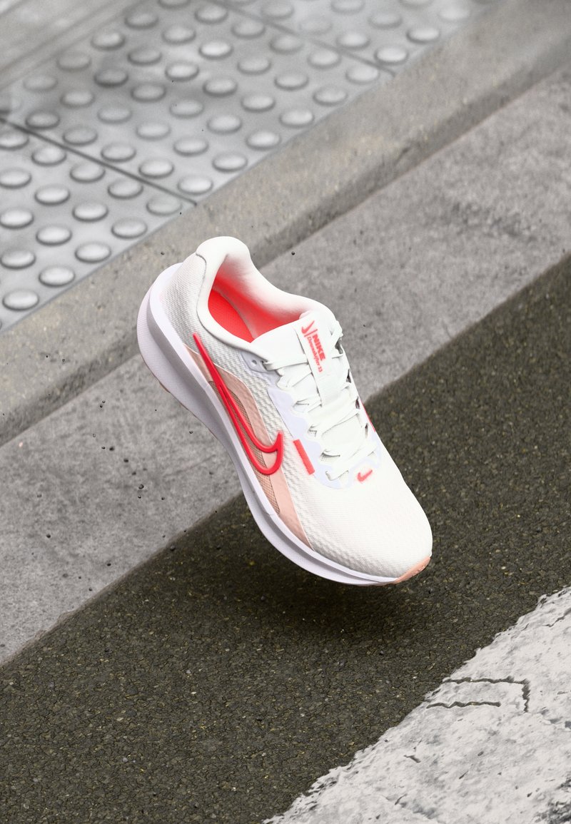 Zapatilla deportiva blanca con un acabado texturizado, acentos en rosa y rojo, suela acolchada y diseño con cordones, sobre un fondo de concreto.