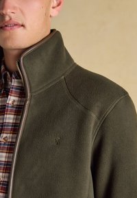 Mann trägt olivgrüne Fleecejacke mit hochgestelltem Kragen über einem rot-weiß-blauen karierten Hemd, beiger Hintergrund.