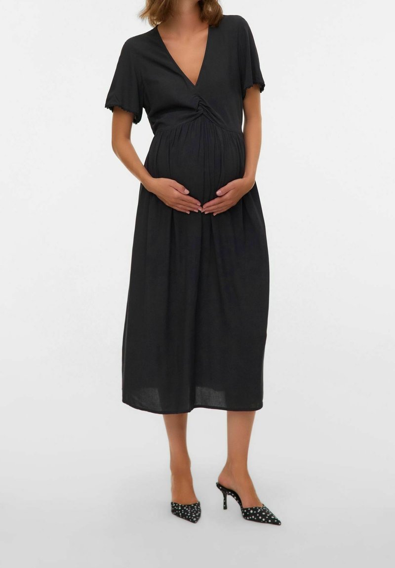 Femme enceinte en robe midi noire à manches courtes, col en V, tenant son ventre, portant des chaussures à talons noirs avec des pois blancs.