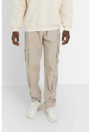 Beige Cargo-Hosen mit mehreren Seitentaschen, aus leichtem Stoff gefertigt. Bieten eine lockere Passform und elastische Bündchen. Kombiniert mit weißen Sneakers.