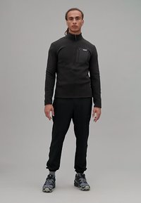 Maglione in pile nero con colletto a zip e tasca sul petto, abbinato a pantaloni neri a gamba affusolata e scarpe da trekking grigie, su uno sfondo neutro.